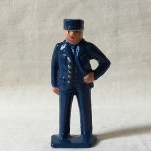 May include: A blue painted metal figurine of a person wearing a blue cap and a blue uniform with gold buttons. The figurine is standing with one hand on their hip.