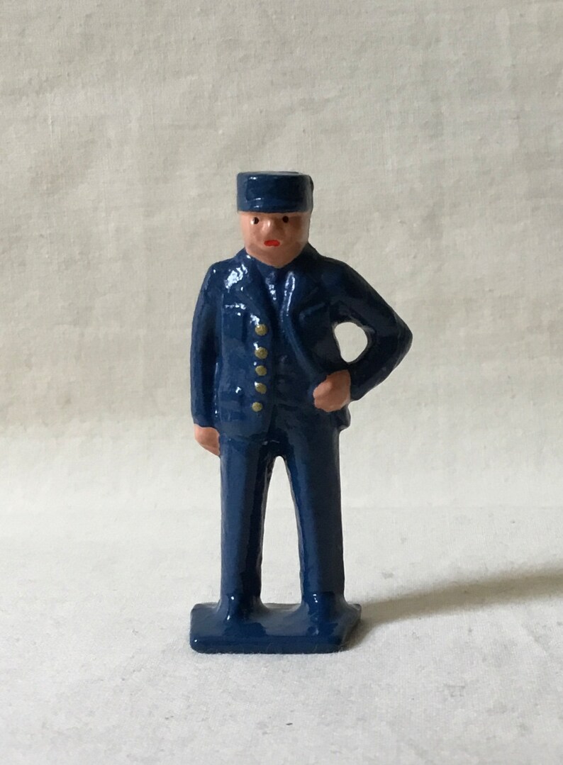 May include: A blue painted metal figurine of a person wearing a blue cap and a blue uniform with gold buttons. The figurine is standing with one hand on their hip.