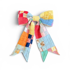 May include: A large hair bow constructed from quilted patchwork fabric featuring a variety of colorful squares in shades of red, yellow, orange, blue, and green. The bow has two long, pointed tails and a central knot, all with visible stitching.