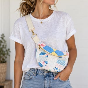 May include: A quilted crossbody bag featuring a patchwork design with blue, yellow, and white floral and geometric patterns. The bag has a gold-tone zipper closure and a wide, adjustable cream-colored canvas strap with gold-tone metal hardware.