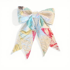 May include: A large, handmade bow made from a patchwork quilt. The bow features a variety of colorful fabric scraps in shades of pink, yellow, blue, and white. The bow is tied in the center with a small loop for attachment.