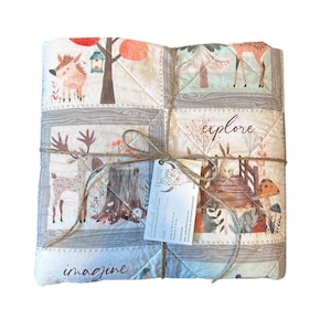 May include: A folded, square quilt with woodland animal and tree illustrations. The quilt is tied with twine and features the words "imagine" and "explore". The color palette includes beige, brown, and green.