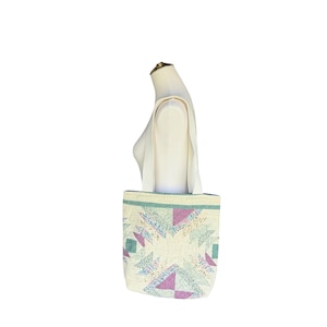 May include: Cream-colored tote bag with a quilted patchwork design. The bag features a geometric pattern in shades of purple, teal, and cream. The bag has a cream-colored strap and is displayed on a mannequin.