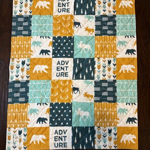 May include: A patchwork quilt featuring squares with various nature-themed designs. The quilt includes images of bears, moose, arrows, and trees in shades of white, teal, mustard yellow, and navy blue. The word "ADVENTURE" is printed on some squares.