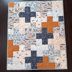 May include: A patchwork baby quilt with a geometric pattern of orange, white, and blue squares. The quilt is made of various fabrics with different patterns, including owls, foxes, and circles.