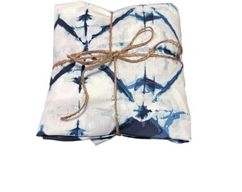 May include: Folded white fabric with a blue tie-dye pattern, tied with twine. The fabric has a repeating design of dark blue shapes against a white background. The twine is wrapped around the fabric and tied in a bow.