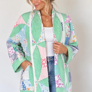 May include: A quilted jacket featuring a patchwork design with various floral and geometric patterns in shades of green, pink, blue, and white. The jacket has long sleeves, an open front with white trim, and two side pockets, constructed from stitched fabric.