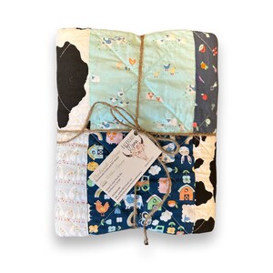 May include: A patchwork quilt with a farm animal theme. The quilt is tied with twine and has a tag that reads "Backroads Charm Quilts Handmade Quilts and Blankets  Chickens & Pigs  Farmhouse Style  Order Online: www.backroadscharmquilts.com  Facebook: Backroads Charm Quilts  @BackroadsCharmQuilts  Paris, IL 61944"