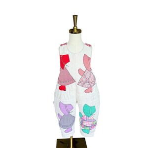 May include: White baby jumpsuit with colorful patchwork cat designs. The sleeveless jumpsuit features quilted fabric and appliqued cat figures in red, pink, purple, and green. The garment is displayed on a mannequin.