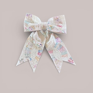 May include: A large, off-white bow with a patchwork design. The bow features a variety of pastel colors, including pink, blue, yellow, and green. The bow is made of a quilted fabric and has long, pointed tails.