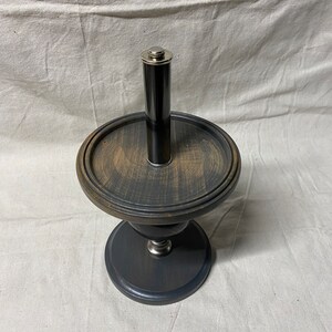 May include: A black wooden pedestal with a round top and a silver metal cylinder in the center. The pedestal has a distressed finish and a dark gray base.