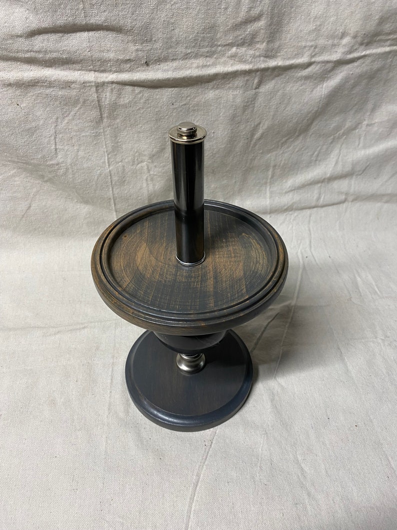 May include: A black wooden pedestal with a round top and a silver metal cylinder in the center. The pedestal has a distressed finish and a dark gray base.