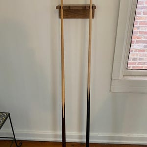 May include: Two pool cues with a wooden wall rack. The cues are light brown with dark brown tips. The rack is made of wood and is mounted on a white wall. The cues are vertical and centered in the image.