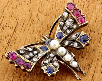Könnte beinhalten: Eine goldene Brosche in Form eines Schmetterlings mit roten und blauen Edelsteinen und Perlen. Die Flügel des Schmetterlings sind mit filigranen Designs verziert.