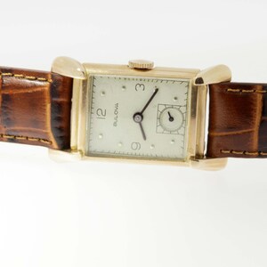 May include: A gold-toned wristwatch with a white face and black hands. The watch has a square face and a brown leather band.
