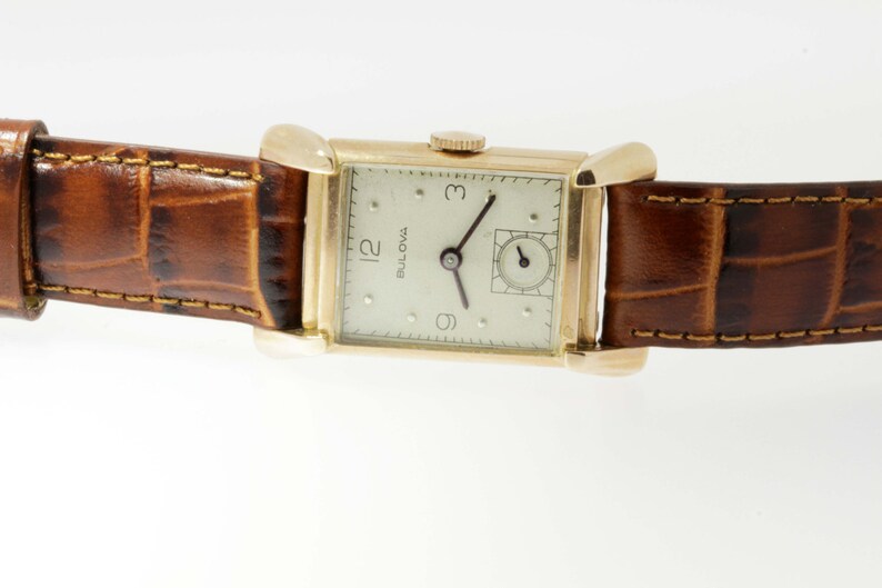 May include: A gold-toned wristwatch with a white face and black hands. The watch has a square face and a brown leather band.