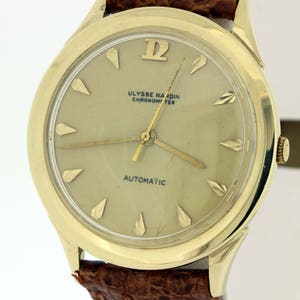 May include: A gold-tone wristwatch with a white face and black hands. The watch face has the text "ULYSSE NARDIN CHRONOMETER AUTOMATIC".