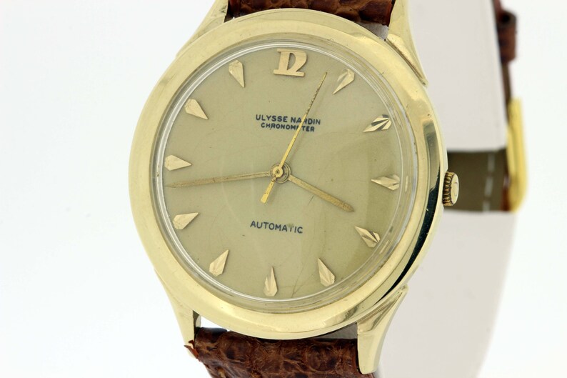 May include: A gold-tone wristwatch with a white face and black hands. The watch face has the text "ULYSSE NARDIN CHRONOMETER AUTOMATIC".