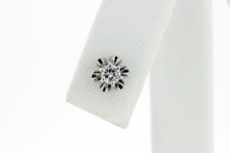 May include: A single diamond earring with a flower-shaped setting. The diamond is clear and round, and the setting is made of white metal.