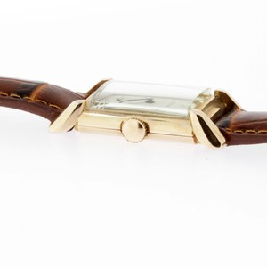 May include: A gold-toned wristwatch with a rectangular face and a brown leather band. The watch has a simple design with a white dial and black hands.