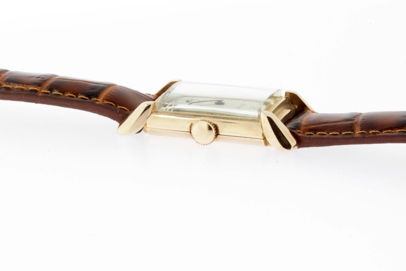 May include: A gold-toned wristwatch with a rectangular face and a brown leather band. The watch has a simple design with a white dial and black hands.