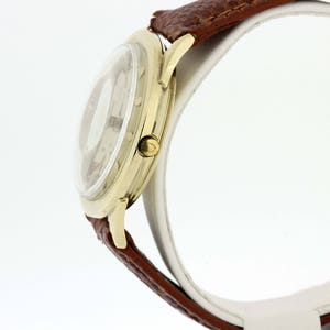 May include: A gold-tone wristwatch with a white face and brown leather strap. The watch is on a white display stand.