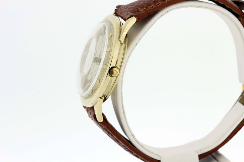 May include: A gold-tone wristwatch with a white face and brown leather strap. The watch is on a white display stand.