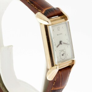 May include: A gold-toned Bulova wristwatch with a white face and black hands. The watch has a rectangular case and a brown leather band.