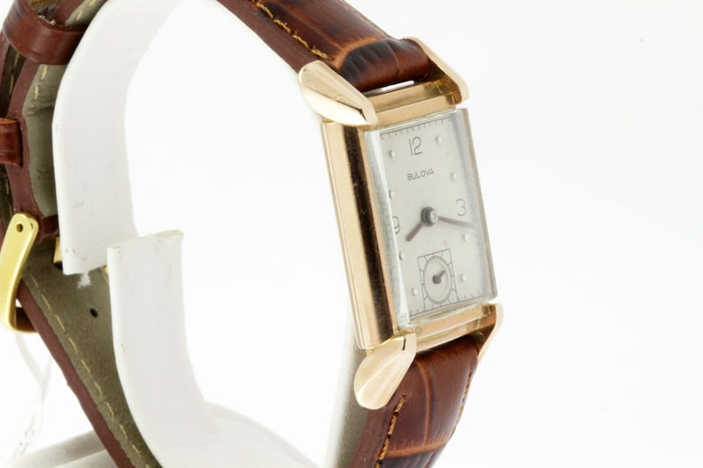 May include: A gold-toned Bulova wristwatch with a white face and black hands. The watch has a rectangular case and a brown leather band.