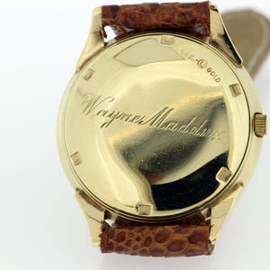 May include: A gold wristwatch with a brown leather strap. The back of the watch has the inscription "18K Gold" and "Wayne Mandabach".