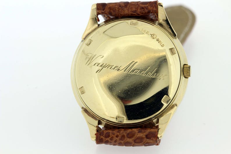 May include: A gold wristwatch with a brown leather strap. The back of the watch has the inscription "18K Gold" and "Wayne Mandabach".