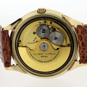 May include: A gold-tone wristwatch with a brown leather strap. The back of the watch is open, revealing the intricate mechanics of the movement. The watch face reads "Ulysse Nardin Swiss Seventeen 17 Jewels Adjusted Temperature".