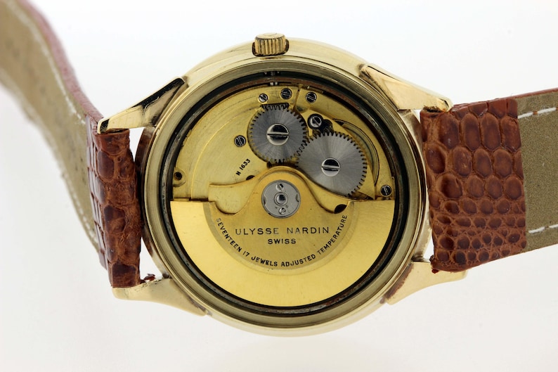 May include: A gold-tone wristwatch with a brown leather strap. The back of the watch is open, revealing the intricate mechanics of the movement. The watch face reads "Ulysse Nardin Swiss Seventeen 17 Jewels Adjusted Temperature".