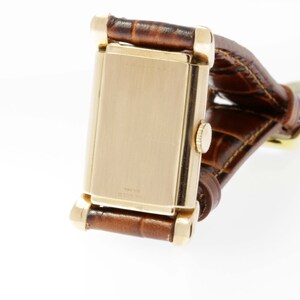 May include: A gold-toned wristwatch with a rectangular face and a brown leather band. The watch has a simple design and is in good condition.