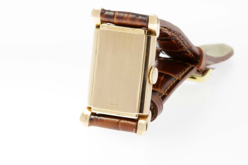 May include: A gold-toned wristwatch with a rectangular face and a brown leather band. The watch has a simple design and is in good condition.