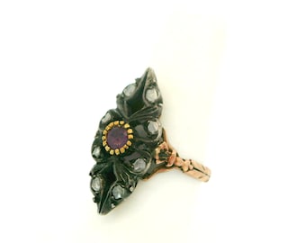 Puede incluir: Anillo antiguo con un engaste oscuro en forma de hoja. Presenta una piedra preciosa púrpura central en un bisel dorado, rodeada de pequeñas piedras transparentes. La banda dorada tiene un diseño ornamentado.