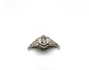 May include: A vintage white gold ring with a central hexagonal setting holding a small, clear gemstone. The ring features intricate filigree detailing and floral patterns on the shoulders, creating an elegant and delicate design.