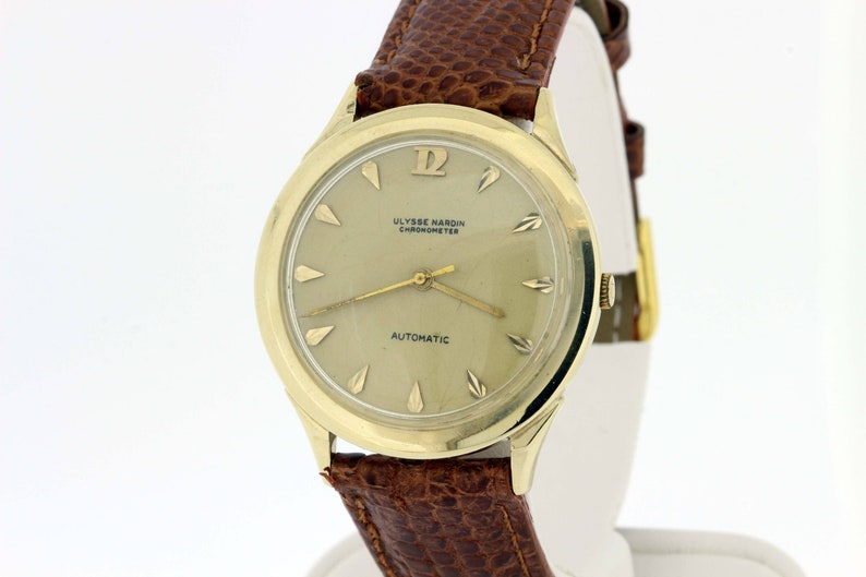 May include: A gold-toned wristwatch with a brown leather strap. The watch face is white with black Roman numeral hour markers and black hands. The watch face also has the text "ULYSSE NARDIN CHRONOMETER AUTOMATIC".