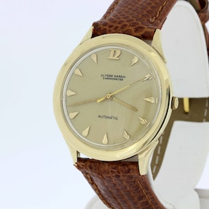 May include: A gold-tone wristwatch with a brown leather strap. The watch face is white with black markings and the text "ULYSSE NARDIN CHRONOMETER AUTOMATIC".
