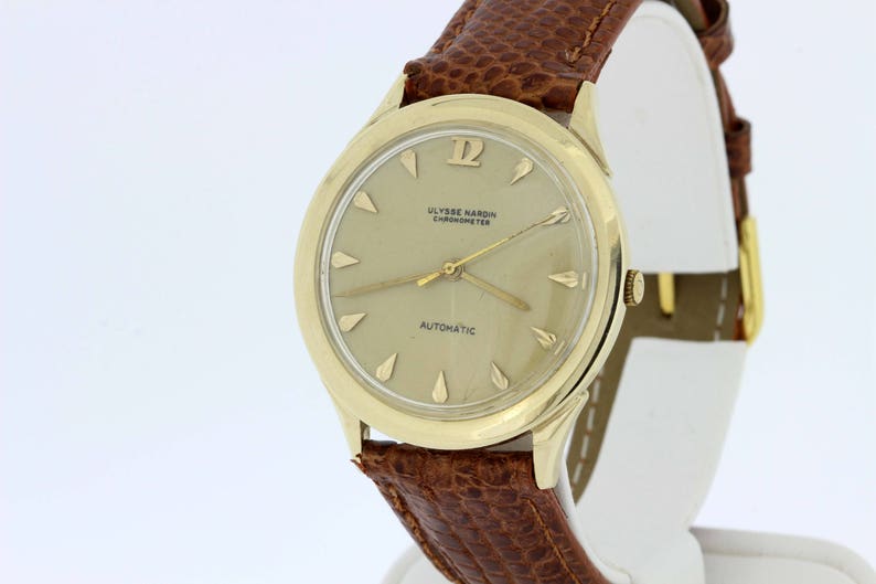 May include: A gold-tone wristwatch with a brown leather strap. The watch face is white with black markings and the text "ULYSSE NARDIN CHRONOMETER AUTOMATIC".