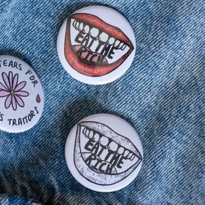 May include: Three white button pins with different designs. The first pin has a pink flower with two red drops of blood and the text "No Tears For Class Traitors". The second and third pins have a red and black mouth with teeth and the text "Eat The Rich".