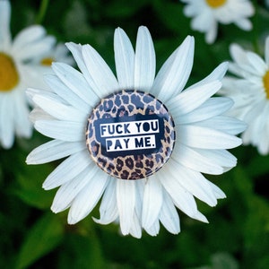 May include: A round button with a leopard print background and the text "FUCK YOU PAY ME" in white, centered on a white daisy. The daisy is surrounded by other daisies and green foliage.