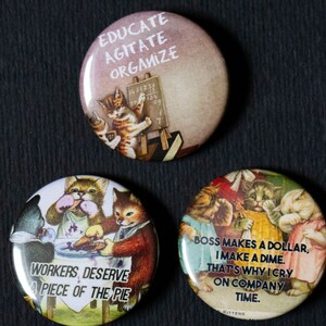 Peut inclure: Trois badges ronds avec des images de chatons. Le premier badge représente un chaton peignant sur un tableau noir avec le texte "Educate Agitate Organize". Le deuxième badge représente des chatons autour d'une table avec une tarte et le texte "Workers Deserve a Piece of the Pie". Le troisième badge représente des chatons en cercle avec le texte "Boss makes a dollar, I make a dime. That's why I cry on company time."