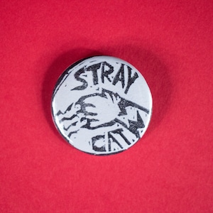 以下が含まれることがあります： 黒文字とグラフィックが描かれた白いボタン。テキストは「Stray Cat」です。
