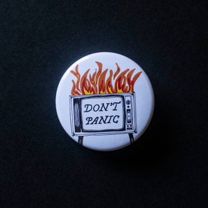 Peut inclure: Bouton blanc avec une illustration en noir et blanc d'un téléviseur en feu avec le texte "DON'T PANIC" sur l'écran.