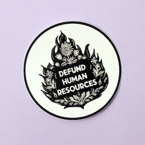 May include: A round sticker with a black and white design. The design features the text "DEFUND HUMAN RESOURCES" within a stylized flame and floral border. The sticker has a white background with a black border, set against a light purple backdrop.