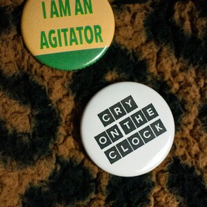 May include: Two round buttons with text. The top button is yellow with green text that says "I am an agitator". The bottom button is white with black text that says "Cry on the clock".