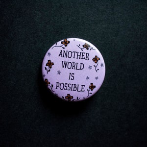 以下が含まれることがあります： 紫色の背景に花柄が描かれた白いボタン。ボタンには「Another World Is Possible.」と書かれています。