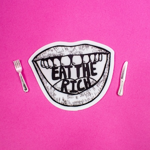 May include: A sticker with a black and white illustration of an open mouth with the text "EAT THE RICH" inside. A silver fork and knife are placed on either side of the sticker against a bright pink background.