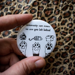 May include: White button with black text and illustrations. The text reads "community care means no one gets left behind". The illustrations include a can of food, a cat, a box, and three cat paws.